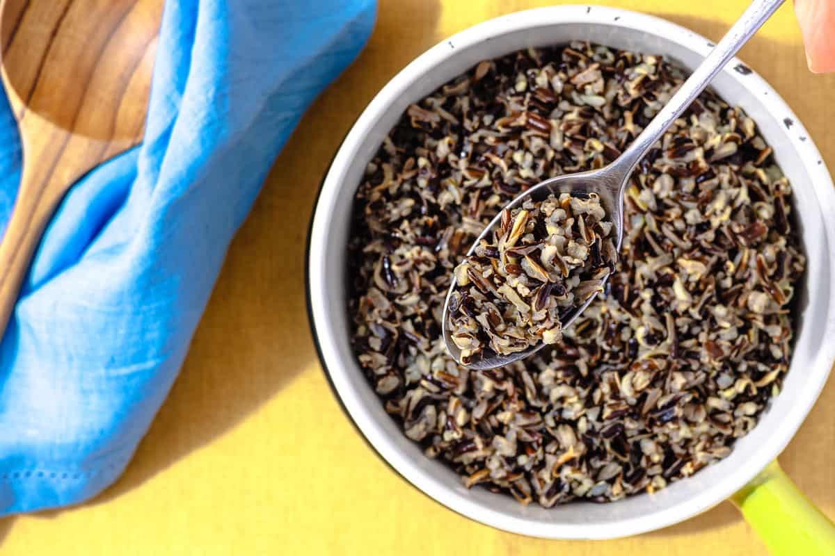 wild rice in a white bowl with a wooden spoon on the side. A metal spoon has a bite full of rice, showing the variety of colors and textures.