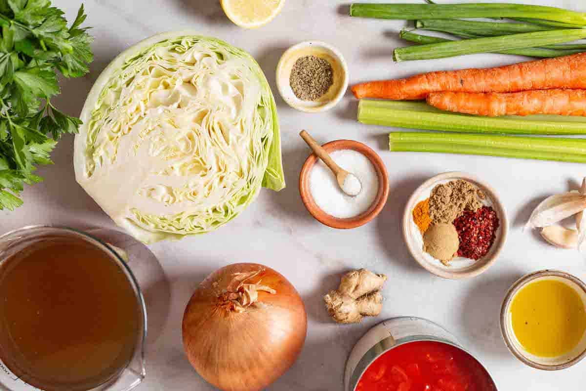 Ingredients for detox cabbage soup including olive oil, onion, garlic, green cabbage, celery, carrots, salt pepper, turmeric, cumin, coriander, Aleppo pepper, diced tomatoes, vegetable broth, parsley, green onions, fresh ginger, and lemon.