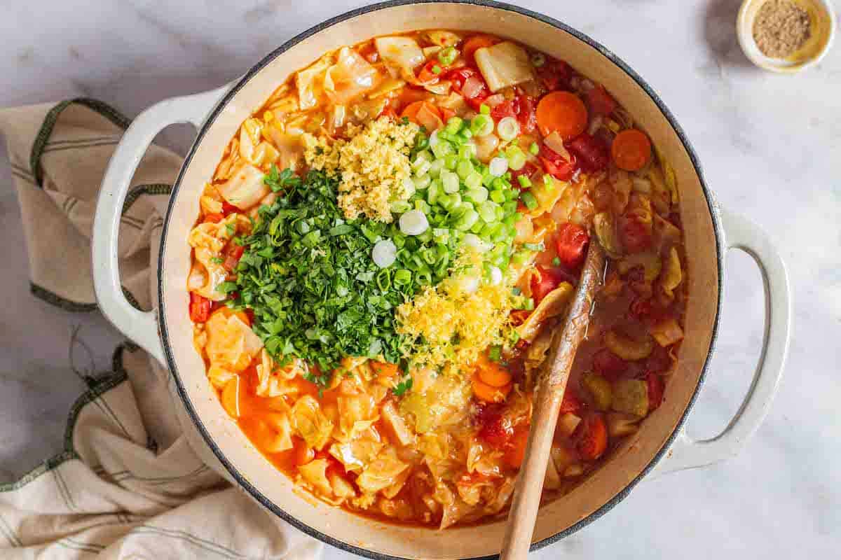 parsley, green onions, ginger, lemon juice and zest added to the pot of detox cabbage soup with a wooden spoon. This is surrounded by a small bowl of pepper, and a cloth napkin.