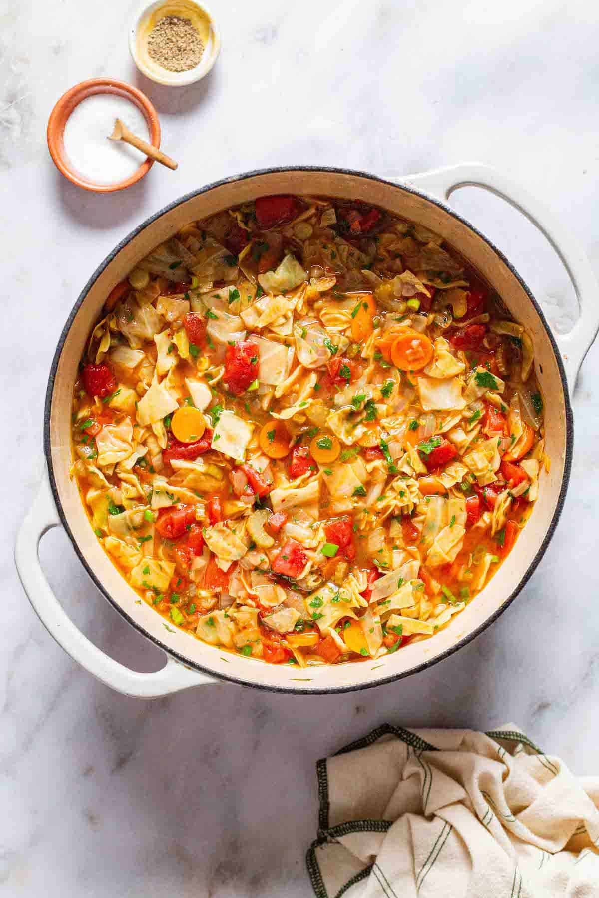 An overhead photo of a pot of detox cabbage soup surrounded by small bowls of salt and pepper and a cloth napkin.