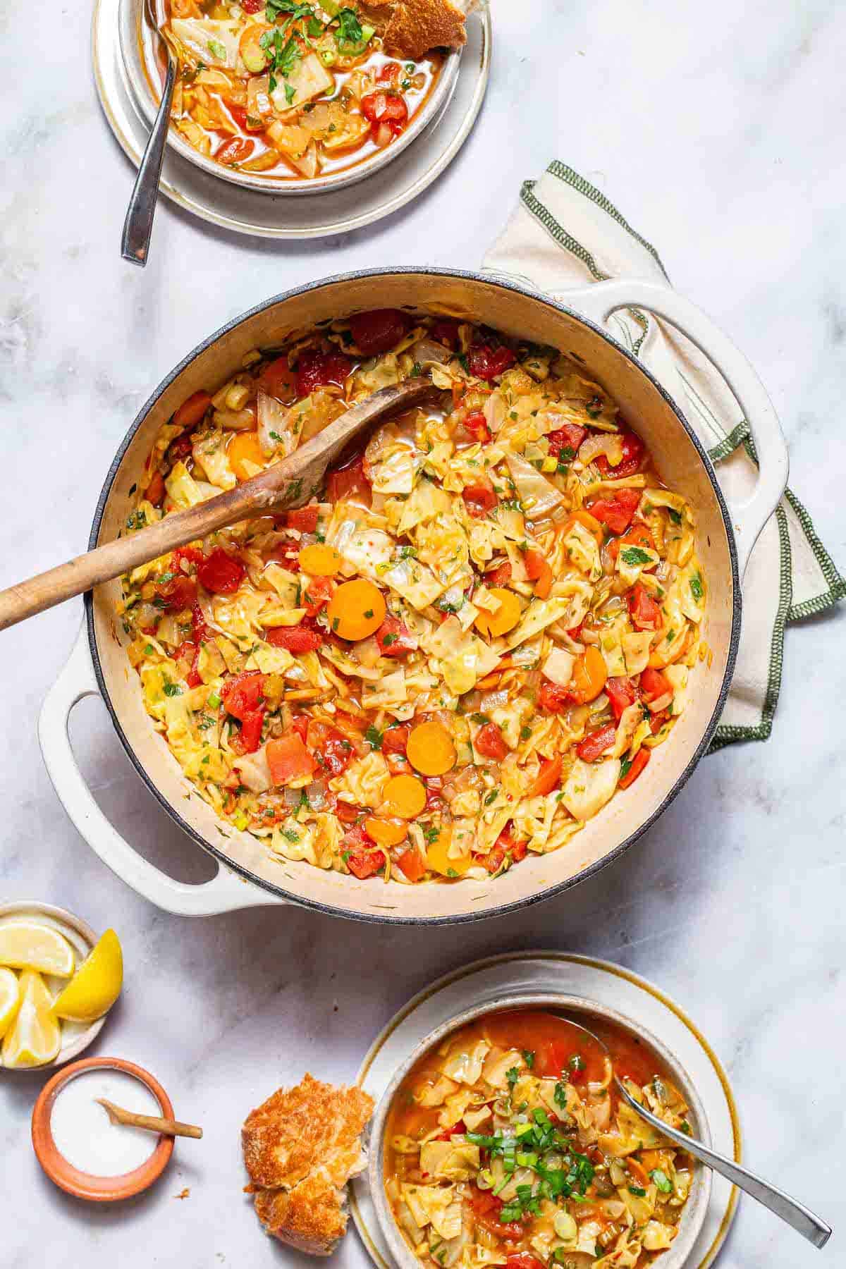 An overhead photo of a pot of detox cabbage soup with a wooden spoon. This is is surrounded by two bowls of the soup with spoons, small bowls up salt and lemon wedges and some pieces of crusty bread.