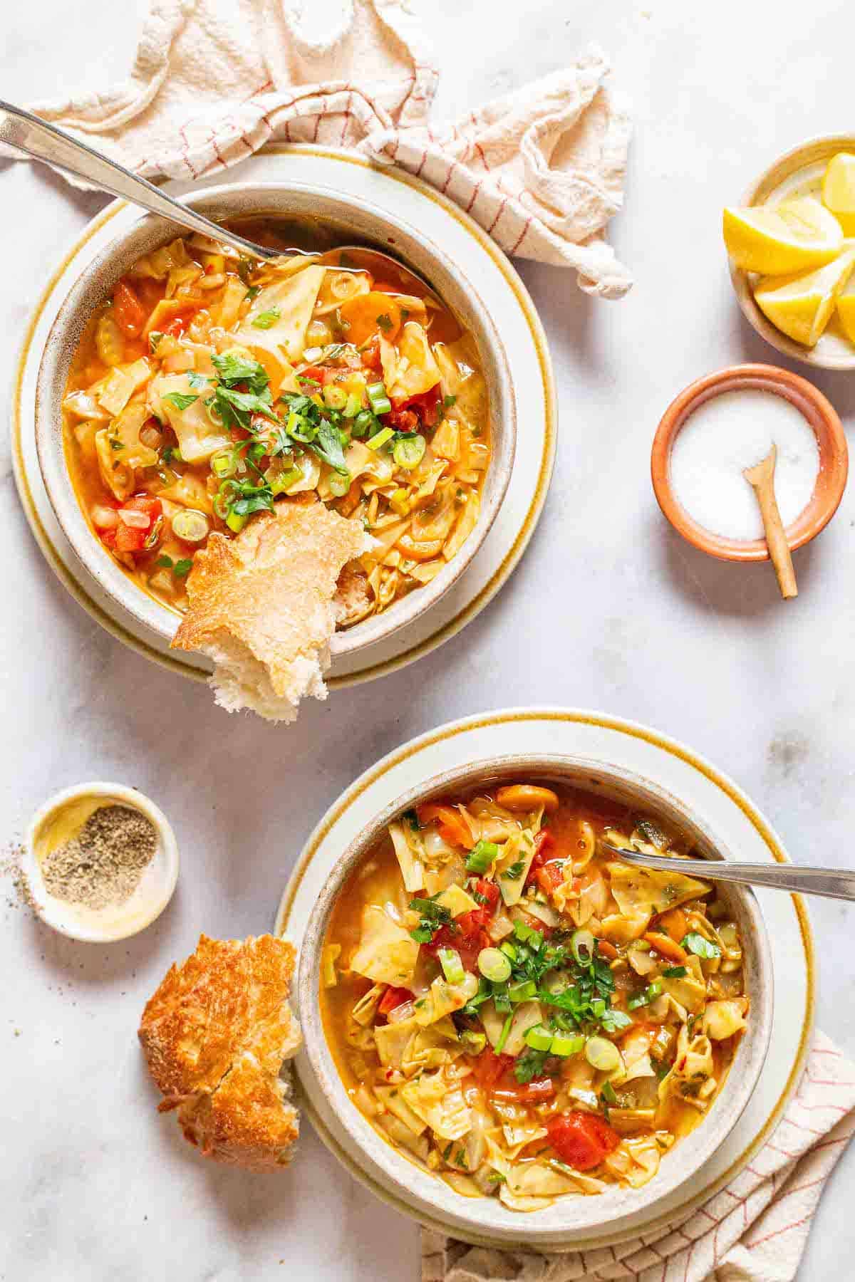 an overhead photo of 2 bowls of detox cabbage soup with spoons surrounded by small bowls of salt, pepper and lemon wedges, slices of crusty bread and cloth napkins.