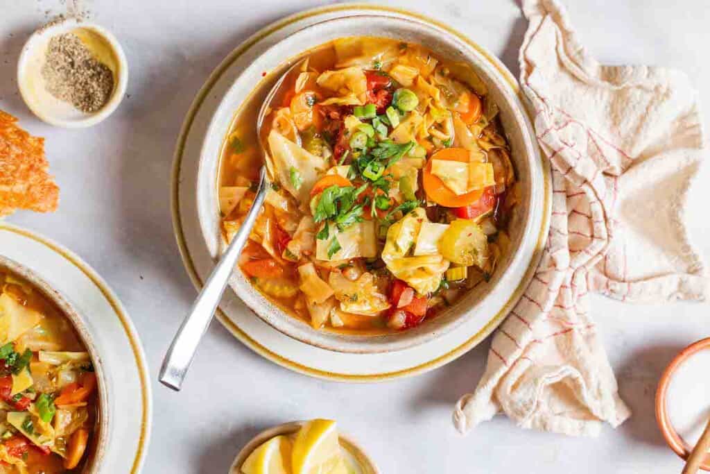 an overhead photo of detox cabbage soup in a bowl with a spoon surrounded by small bowls of salt, pepper and lemon wedges and a cloth napkin.