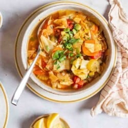 an overhead photo of detox cabbage soup in a bowl with a spoon surrounded by small bowls of salt, pepper and lemon wedges and a cloth napkin.