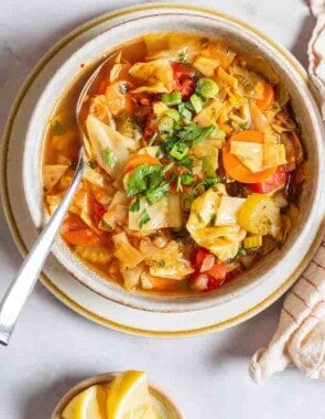 an overhead photo of detox cabbage soup in a bowl with a spoon surrounded by small bowls of salt, pepper and lemon wedges and a cloth napkin.