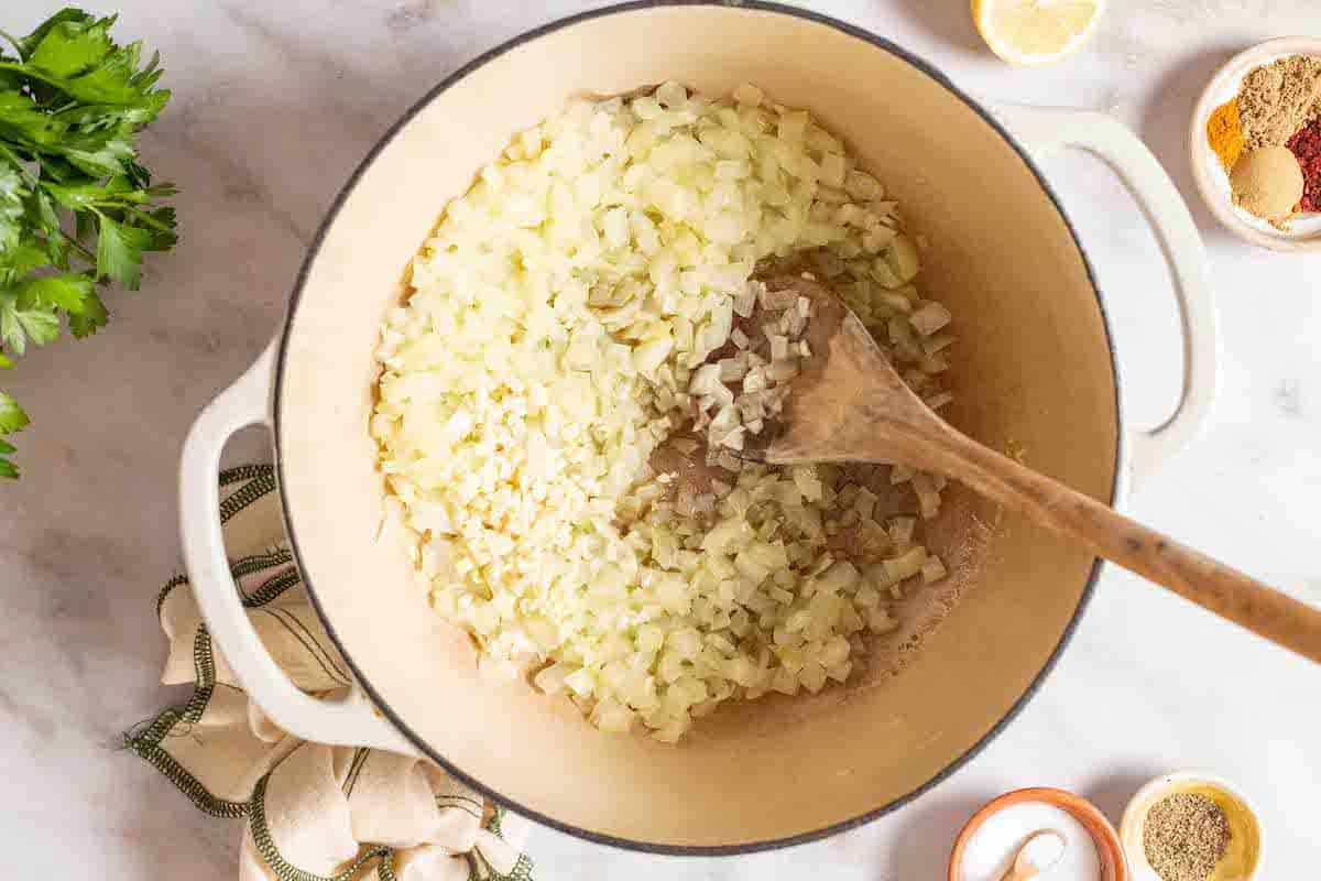 Chopped onion and minced garlic being sauteed in a pot with a wooden spoon. This is surrounded by a lemon half, a bunch of parsley, small bowls of various spices, salt and pepper, and a cloth napkin.