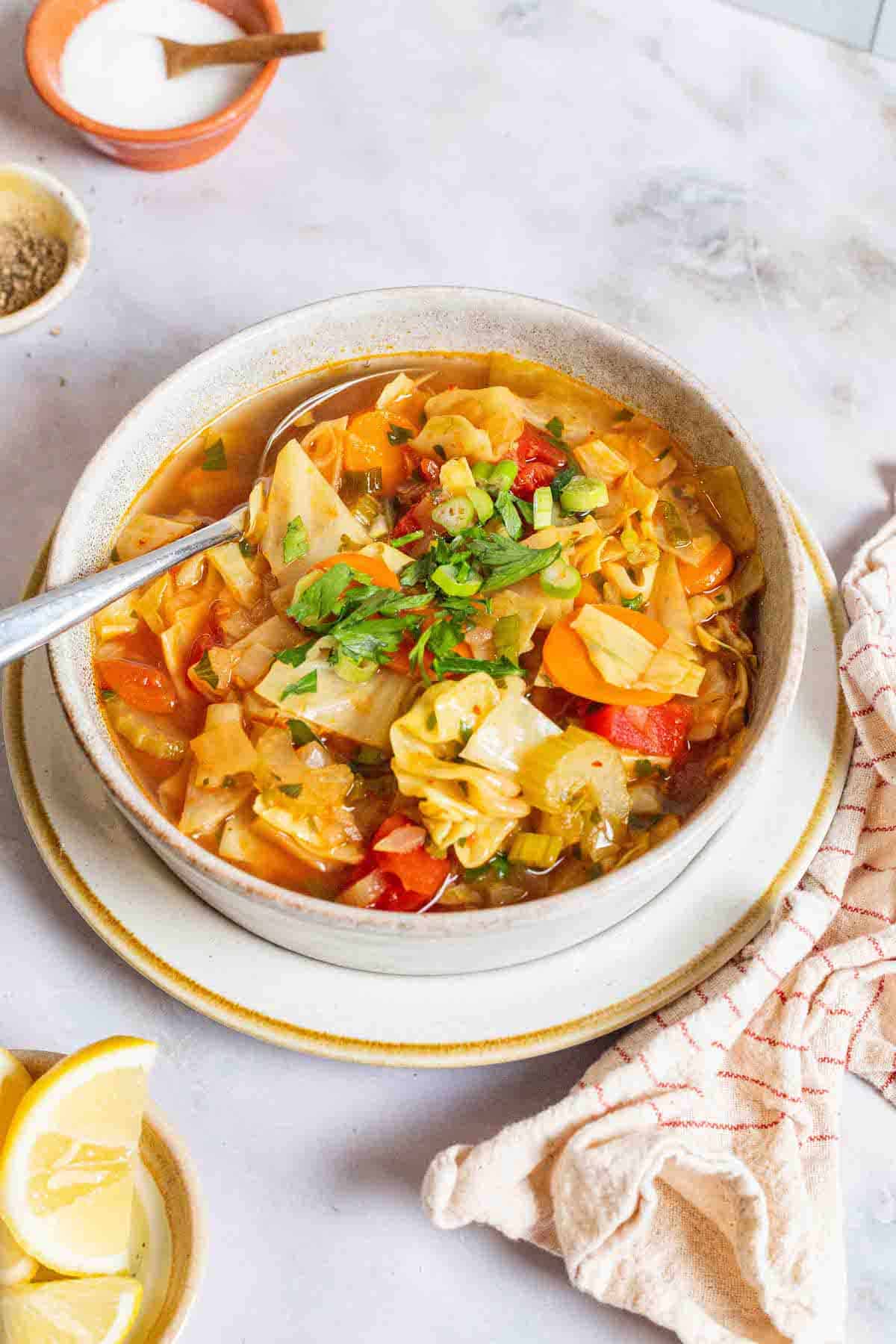 detox cabbage soup in a bowl with a spoon surrounded by small bowls of salt, pepper and lemon wedges and a cloth napkin.