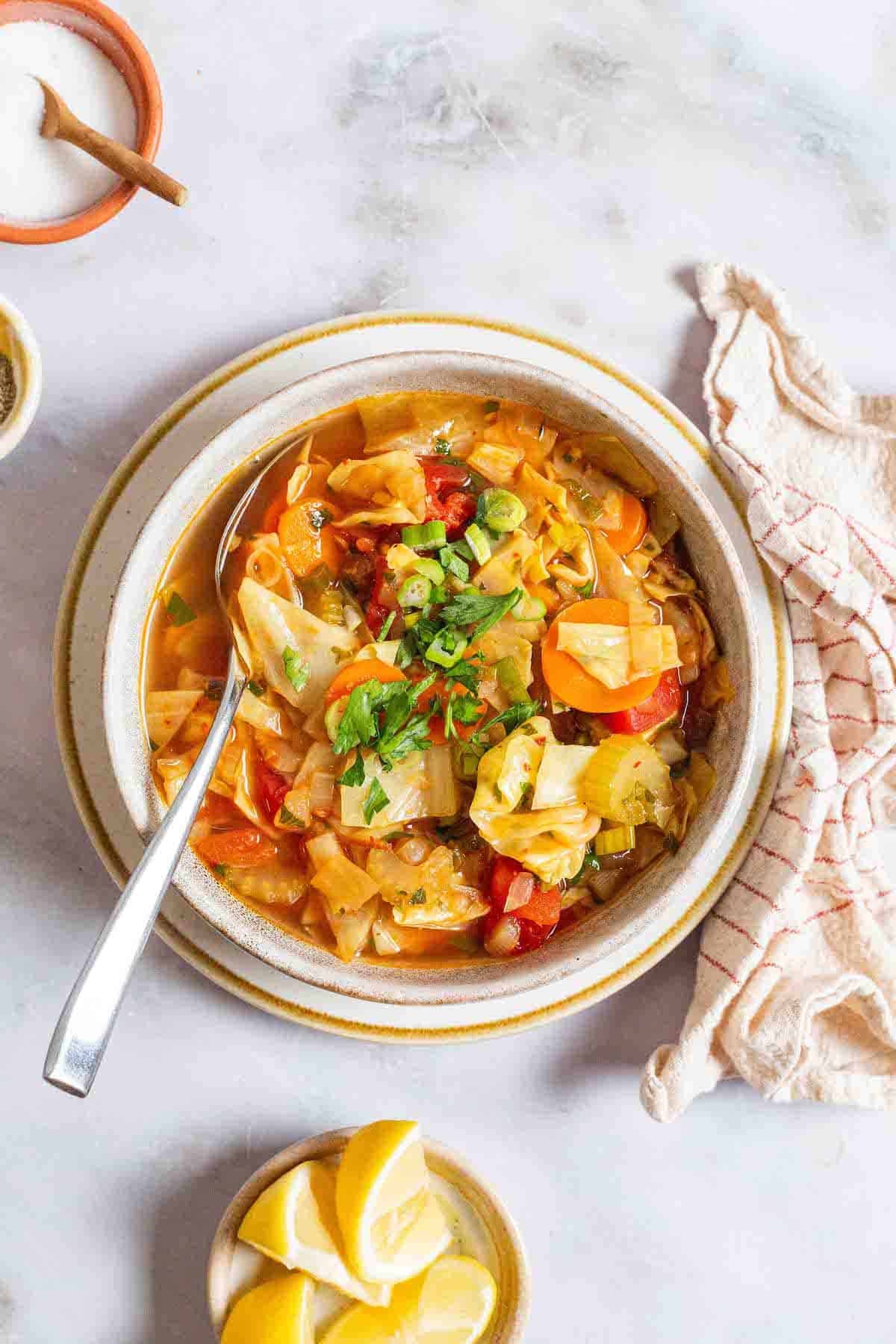 an overhead photo of detox cabbage soup in a bowl with a spoon surrounded by small bowls of salt and lemon wedges and a cloth napkin.