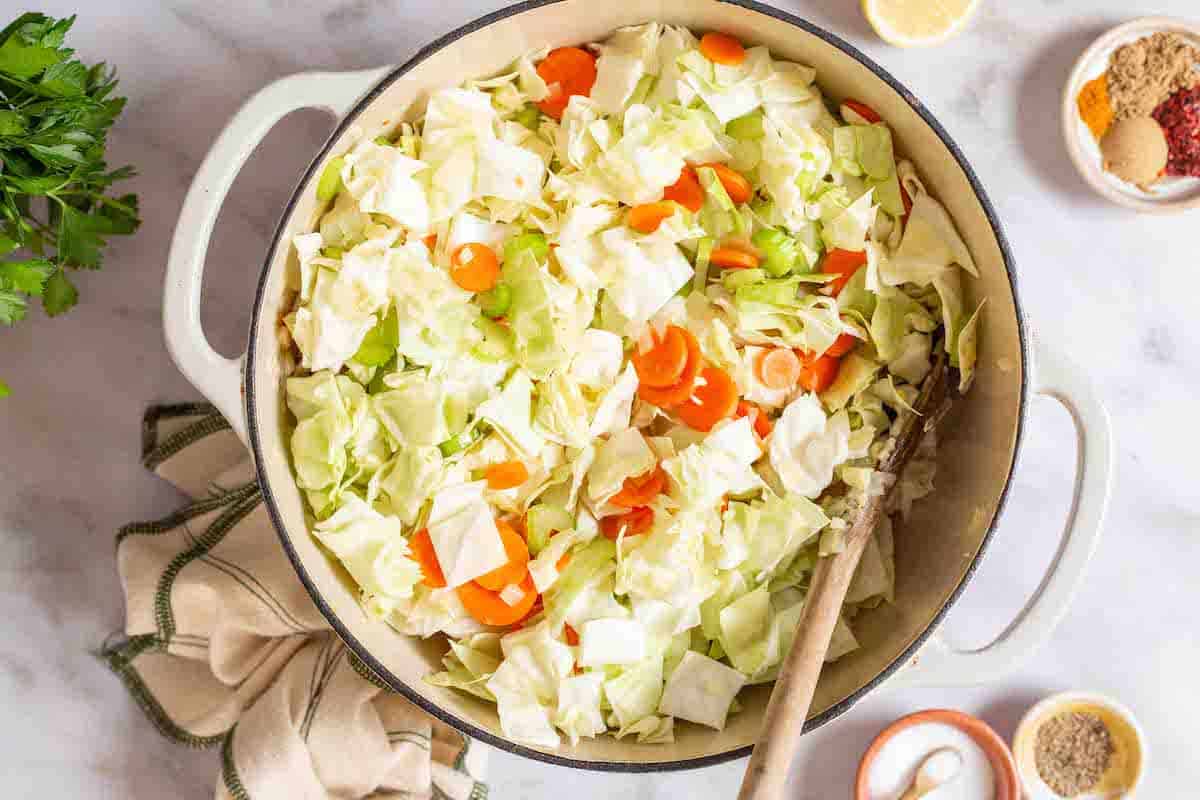cabbage, celery and carrots being softened in a pot with a wooden spoon. This is surrounded by a lemon half, a bunch of parsley, small bowls of various spices, salt and pepper, and a cloth napkin.