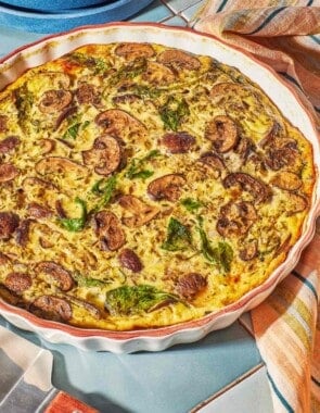 The baked crustless quiche recipe in a baking dish next to a serving utensil, a kitchen towel and a stack of 2 plates.
