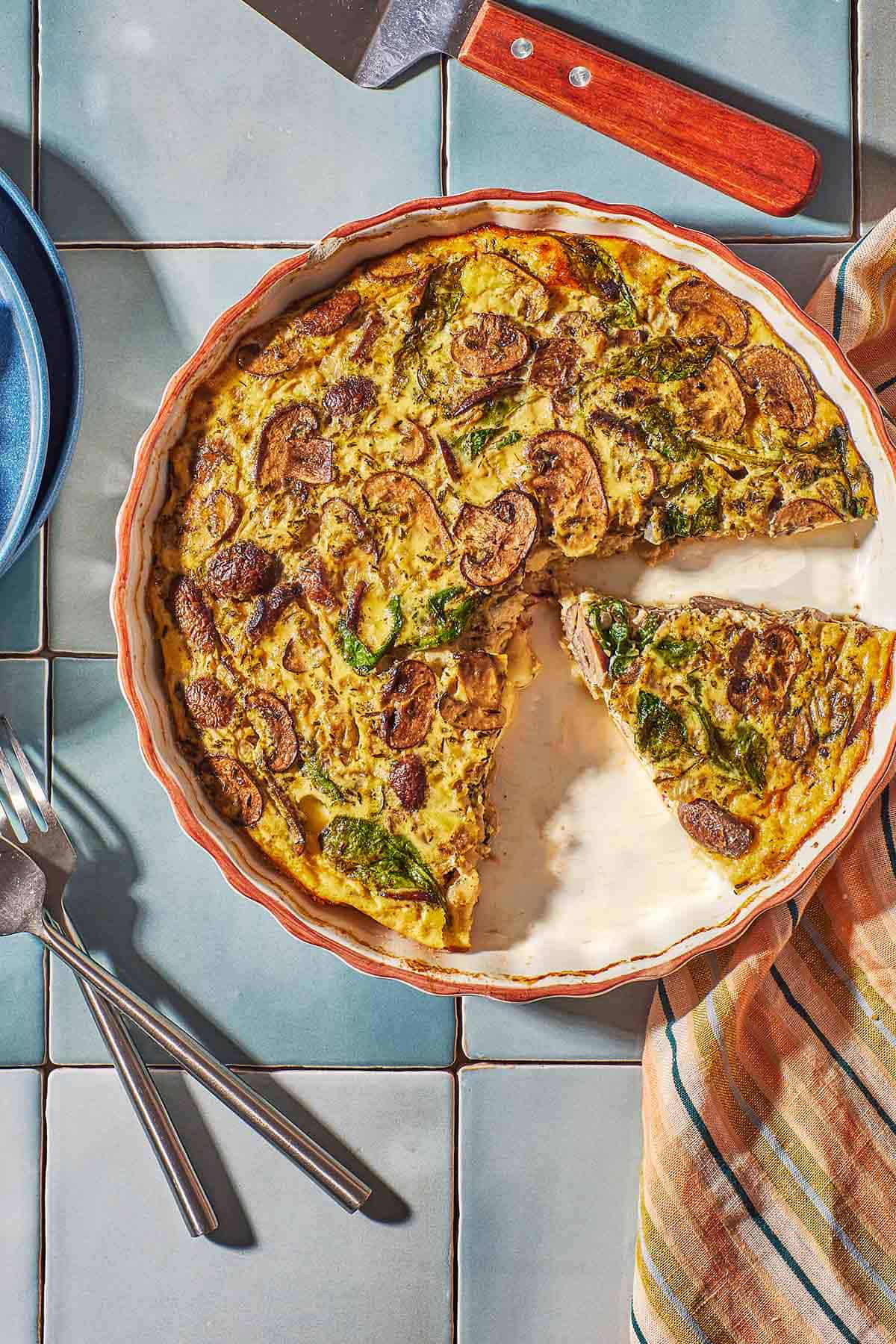 Spinach and mushroom crustless quiche on a tile countertop. Slices of the quiche are missing. 