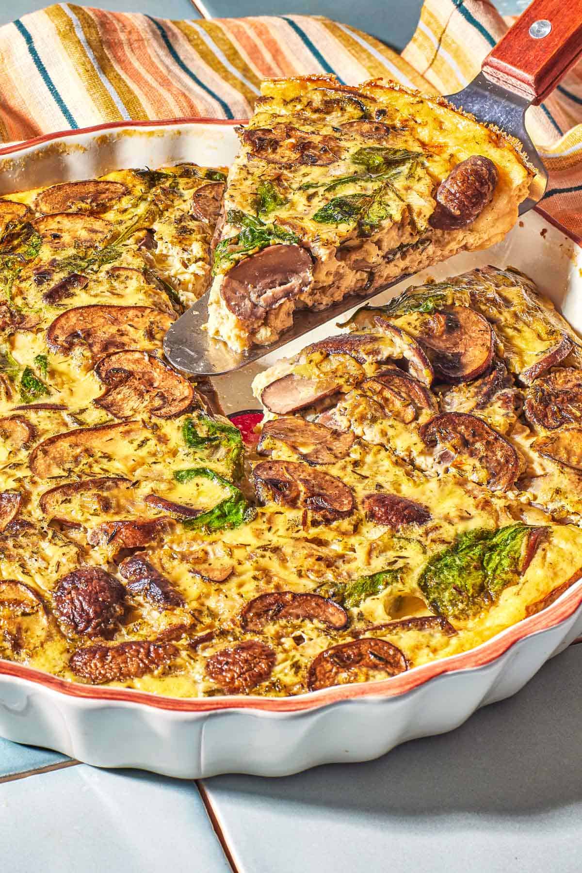 Crustless quiche stuffed with spinach and mushrooms in a quiche plate with a piece of quiche being lifted out of the pan. 