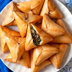 An overhead photo of several Fatayer Spinach and Onion Hand Pies on a serving plate next to a blue cloth napkin. The fatayer on the top is cut in half, exposing the filling inside.