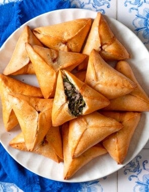 An overhead photo of several Fatayer Spinach and Onion Hand Pies on a serving plate next to a blue cloth napkin. The fatayer on the top is cut in half, exposing the filling inside.