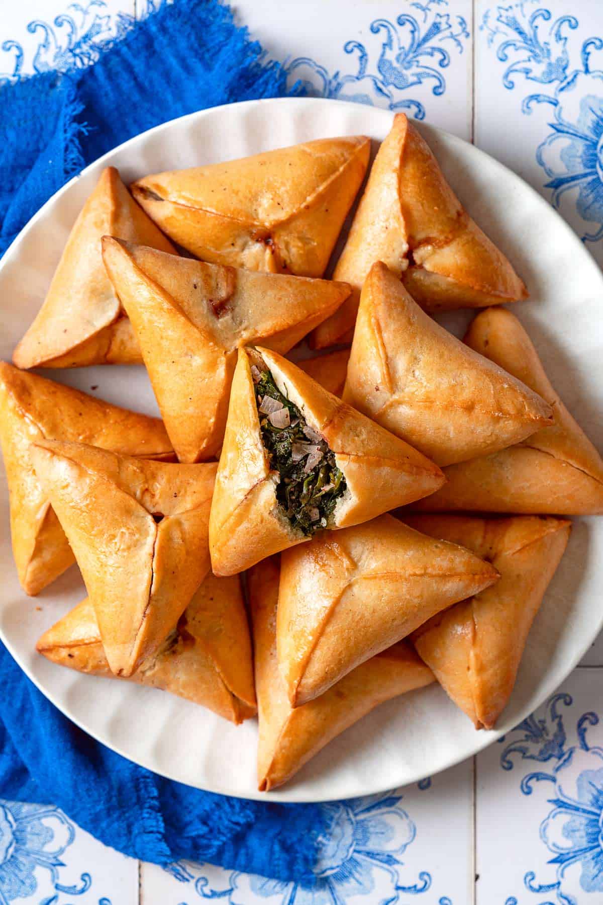 An overhead photo of several Fatayer Spinach and Onion Hand Pies on a serving plate next to a blue cloth napkin. The fatayer on the top is cut in half, exposing the filling inside.