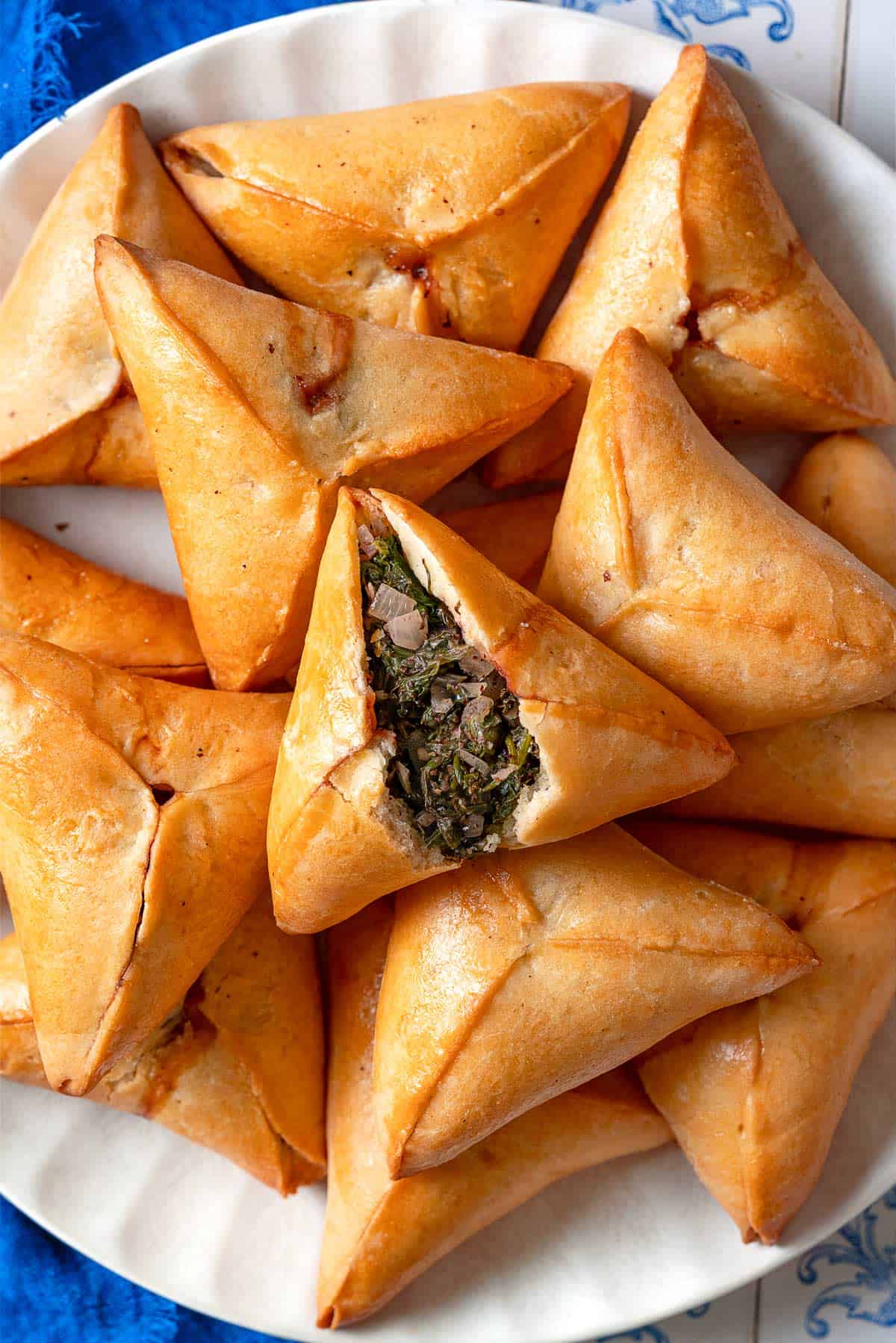 A close up of several Fatayer Spinach and Onion Hand Pies on a serving plate next to a blue cloth napkin. The fatayer on the top is cut in half, exposing the filling inside.