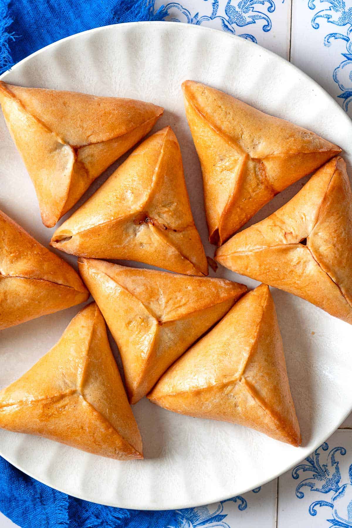 An overhead photo of several Fatayer Spinach and Onion Hand Pies on a serving plate next to a blue cloth napkin.