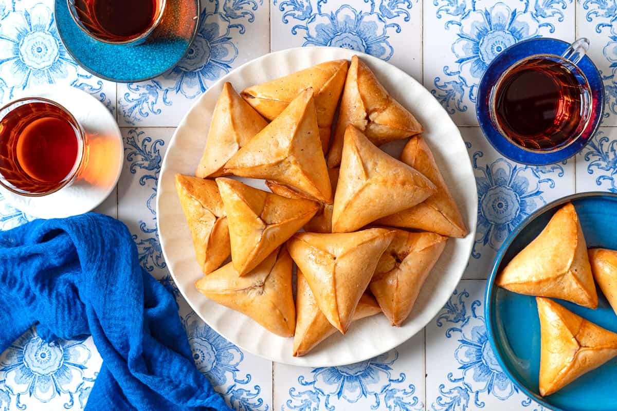 An overhead photo of several Fatayer Spinach and Onion Hand Pies on a serving plate next to a blue cloth napkin, three cups of tea, and a plate of 3 more of the hand pies.