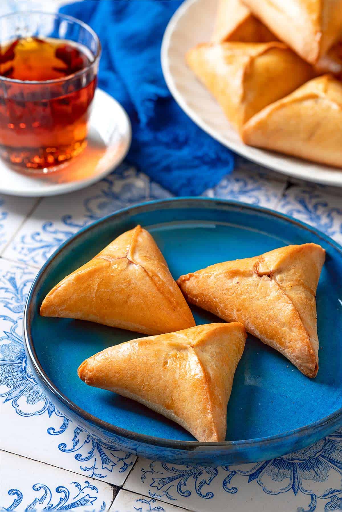 A close up of 23 Fatayer Spinach and Onion Hand Pies on a plate in front of a cup of tea, a cloth napkin and a serving plate with more of the hand pies.