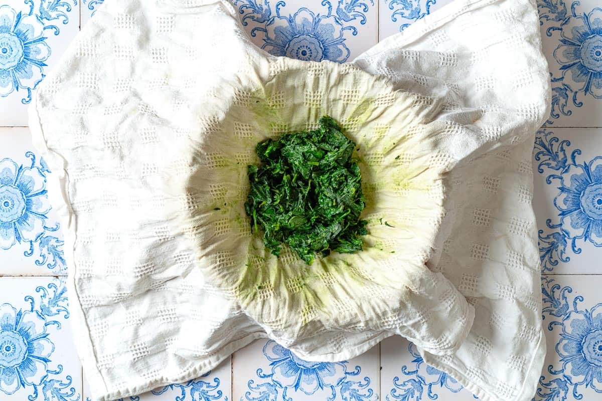 Wilted spinach on a hand towel draped over a bowl after the excess liquid has been squeezed out.