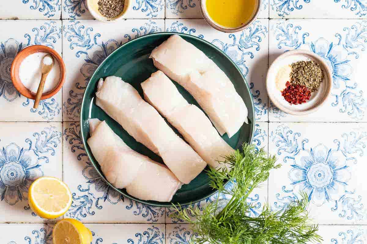 Ingredients for baked halibut including olive oil, 4 pieces of halibut fillet, salt, pepper, Italian seasoning, garlic powder, Aleppo pepper, a lemon and fresh dill.