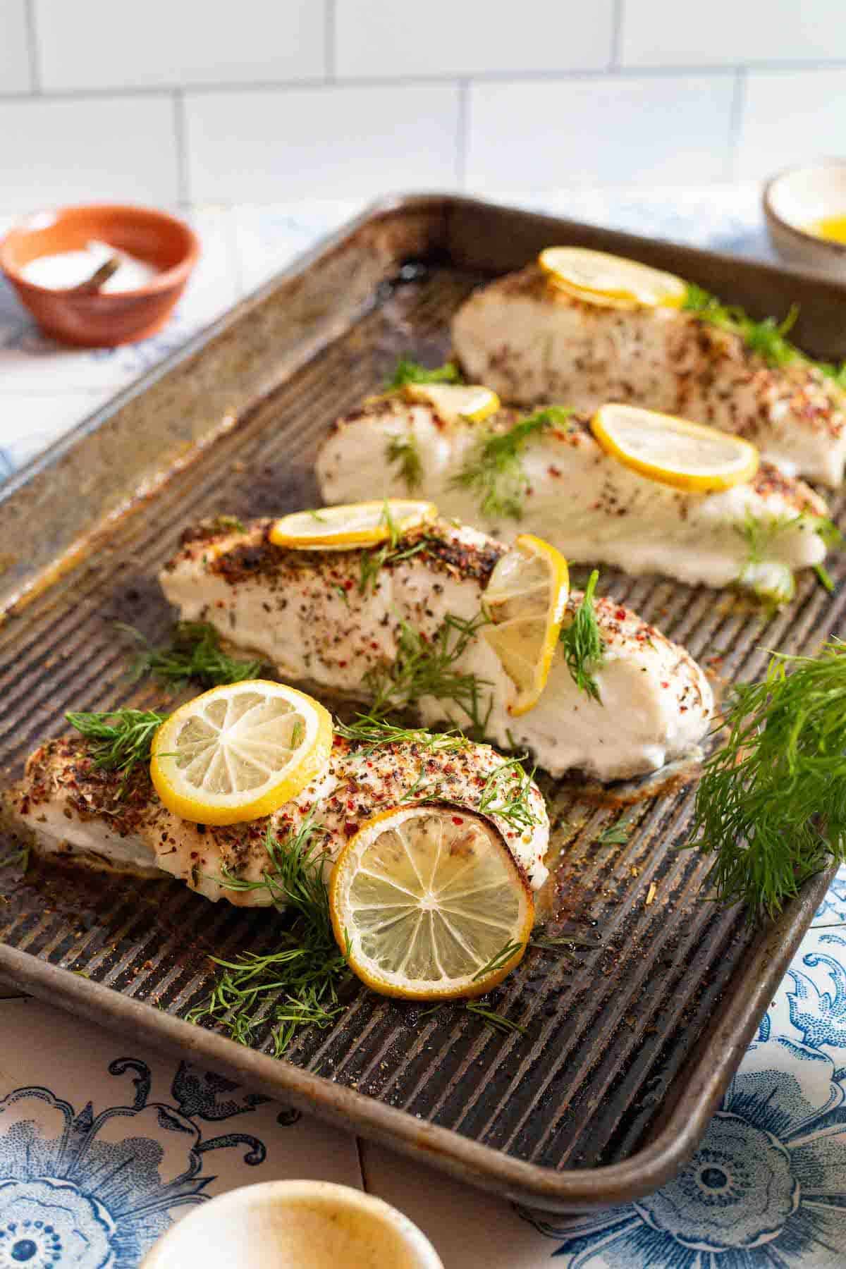 4 baked halibut fillets garnished with lemon and dill on a baking sheet surrounded by small bowls of salt, olive oil and other seasonings.