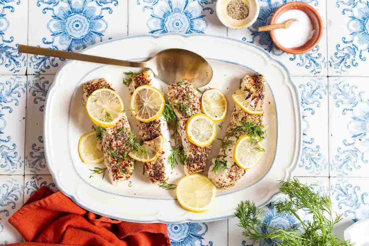 an overhead photo of 4 baked halibut fillets garnished with lemon and dill on a serving platter with a spoon. Surrounding the platter are fresh dill and small bowls of seasonings and salt.