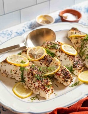 4 baked halibut fillets garnished with lemon and dill on a serving platter with a spoon. In the background, there are small bowls of seasonings and salt.