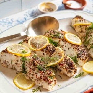 4 baked halibut fillets garnished with lemon and dill on a serving platter with a spoon. In the background, there are small bowls of seasonings and salt.
