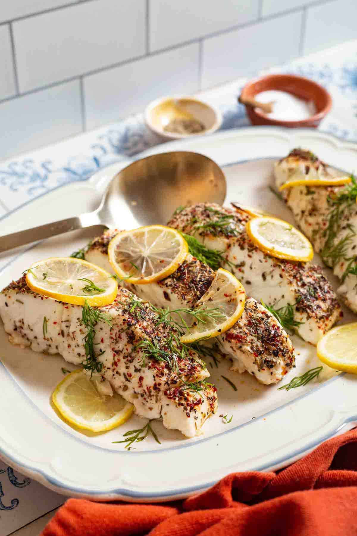 4 baked halibut fillets garnished with lemon and dill on a serving platter with a spoon. In the background, there are small bowls of seasonings and salt.