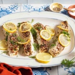 4 baked halibut fillets garnished with lemon and dill on a serving platter. In the background, there are small bowls of seasonings and salt.