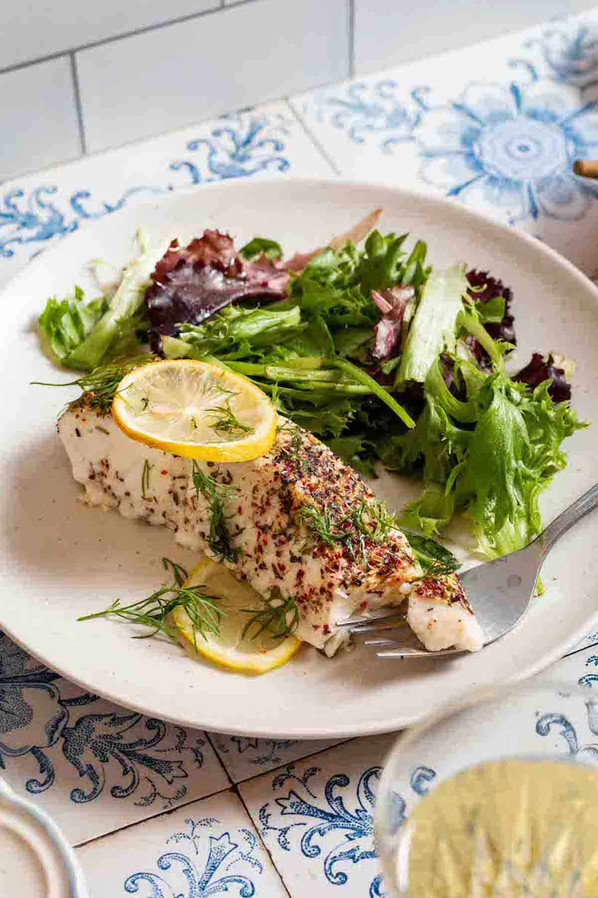 A baked halibut fillet garnished with lemon and dill on a plate with a salad and a fork with a bite of halibut on it.