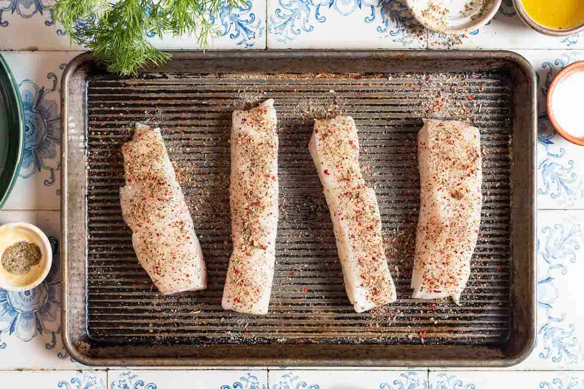 4 seasoned, but unbaked halibut fillets on a baking sheet surrounded by dill and bowls of olive oil, salt, pepper and other seasonings.