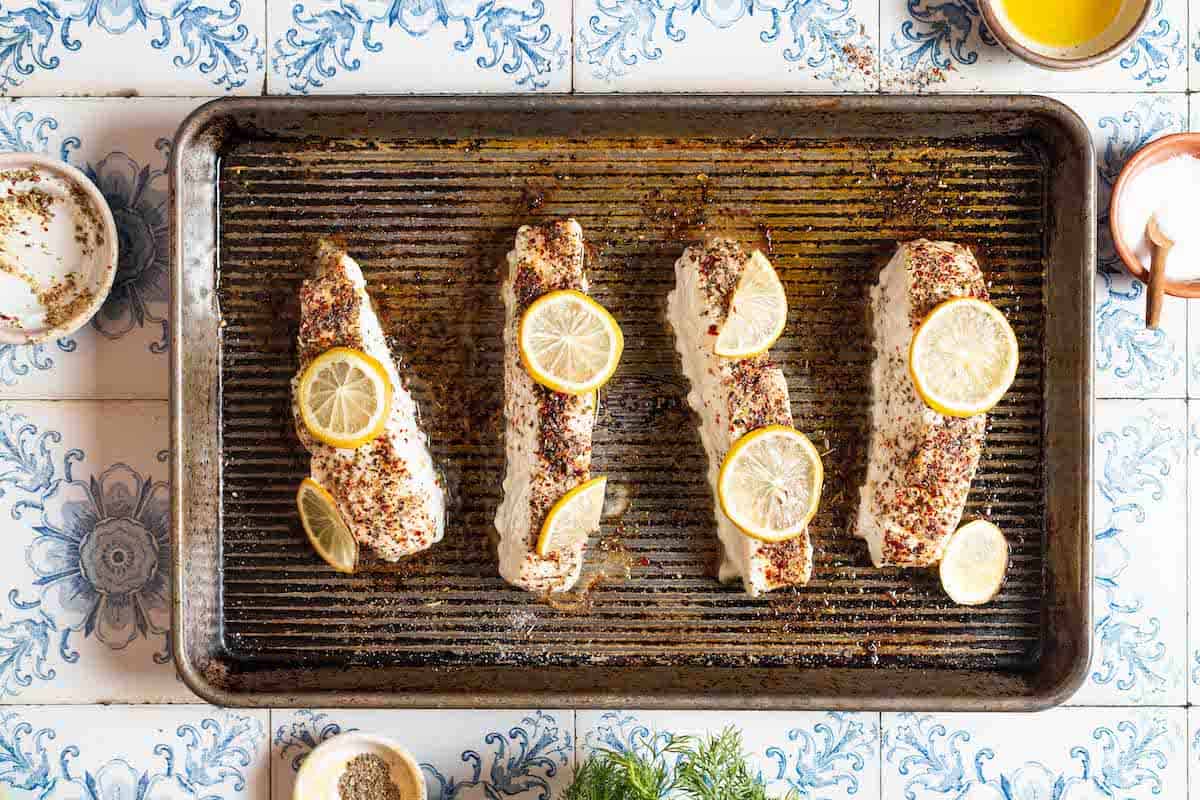 4 baked halibut fillets topped with lemon slices on a baking sheet surrounded by bowls of olive oil, salt, pepper and other seasonings.