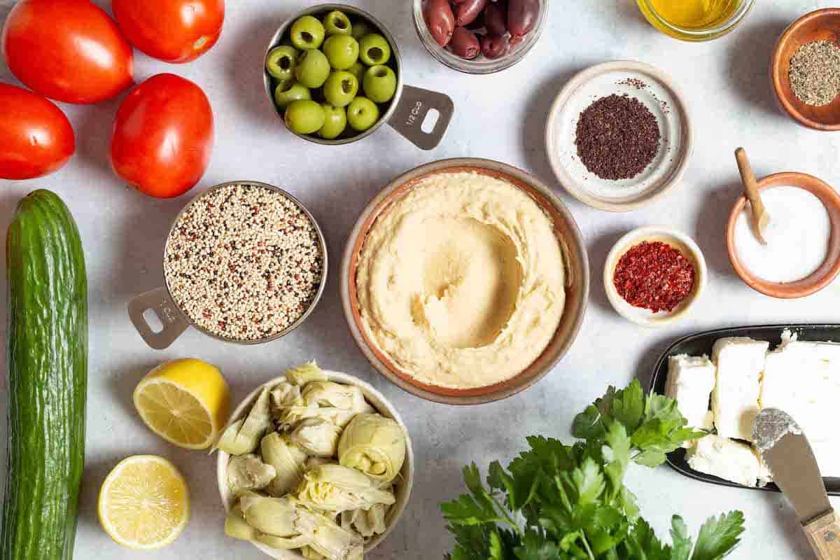 ingredients for mediterranean bowl with quinoa including quinoa, salt, hummus, tomatoes, cucumber, lemon, kalamata olives, green olives, artichoke hearts, feta cheese, parsley, olive oil, Aleppo pepper and sumac.