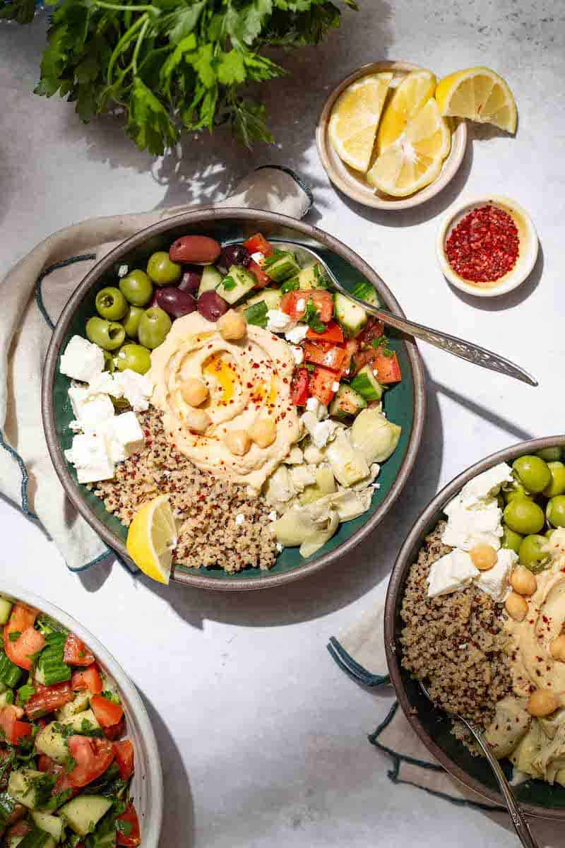 An overhead photo of 2 mediterranean bowls with quinoa with forks in them. These are surrounded by a bunch of parsley, a bowl of lemon slice, a bowl of aleppo pepper, and the Mediterranean cucumber tomato salad.