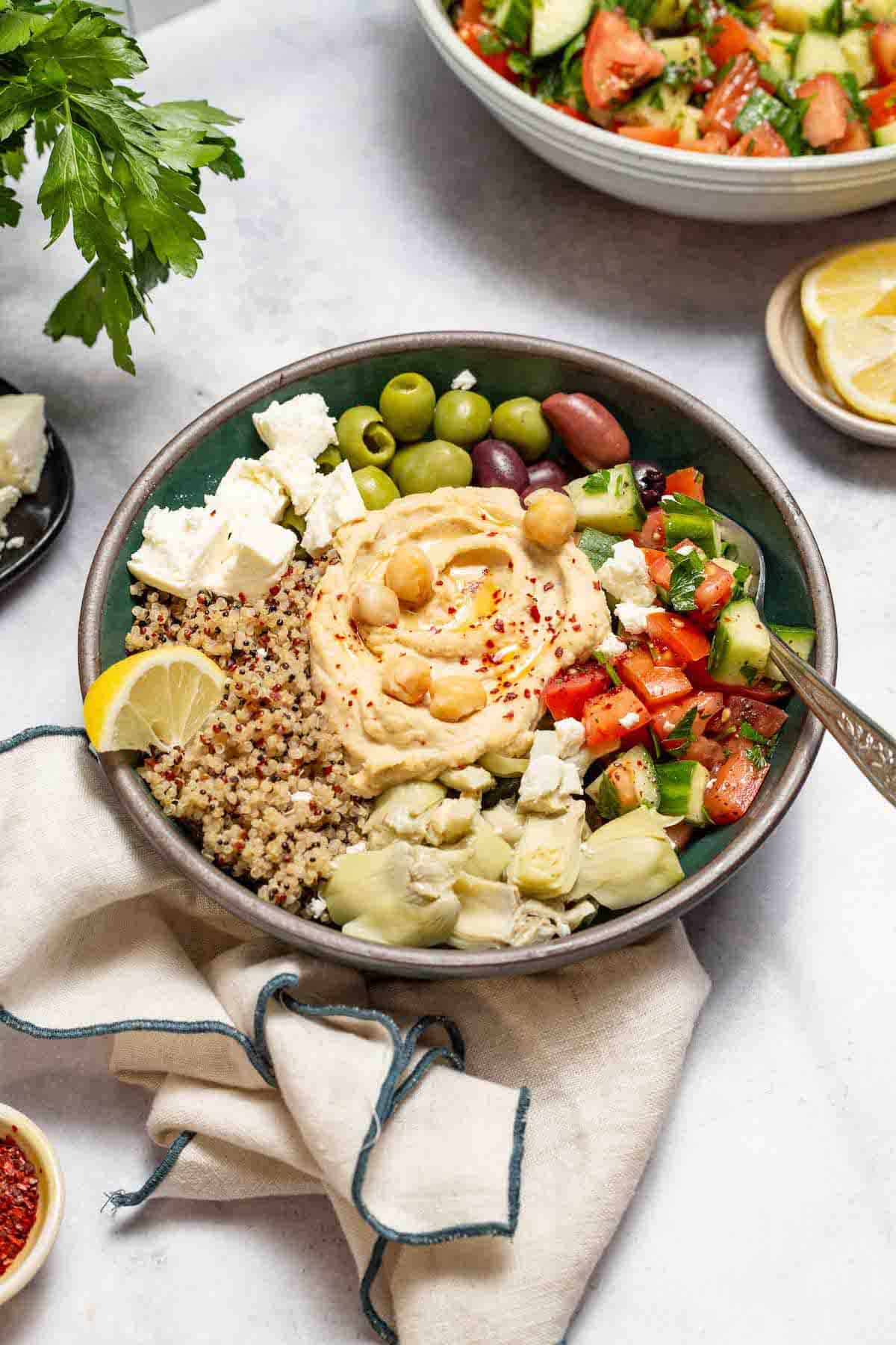 A mediterranean bowl with quinoa with a forks in it surrounded by a cloth napkin, parsley, a bowl of lemon slices, and the mediterranean cucumber tomato salad.
