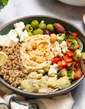 A mediterranean bowl with quinoa with a fork in it. This is surrounded by bowls of lemon slices and feta cheese.