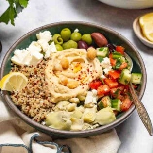 A mediterranean bowl with quinoa with a fork in it. This is surrounded by bowls of lemon slices and feta cheese.