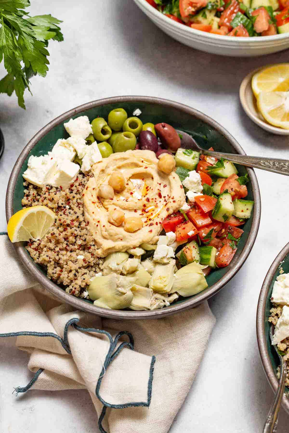 A mediterranean bowl with quinoa with a fork in it next to a cloth napkin, parsley, a Mediterranean cucumber salad, and a bowl of lemon slices.