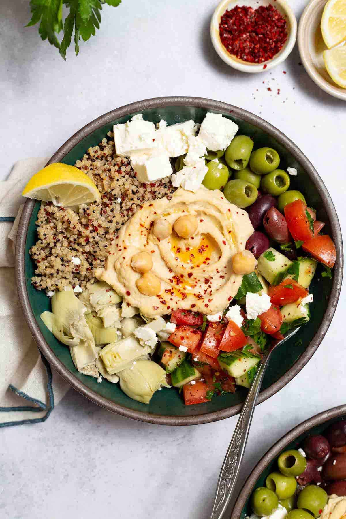 An overhead photo of a mediterranean bowl with quinoa with a fork in it. This is surrounded by a bowl of lemon slices, a bowl of aleppo pepper and another Mediterranean bowl with quinoa.