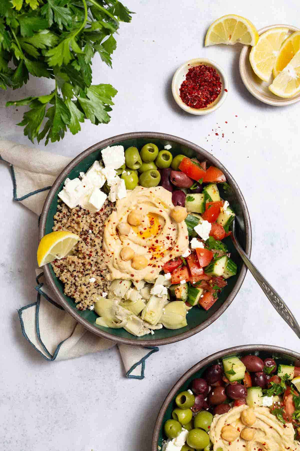 An overhead photo of 2 mediterranean bowls with quinoa, one with a fork in it. These are surrounded by a bunch of parsley, and bowls of lemon slices and aleppo pepper.
