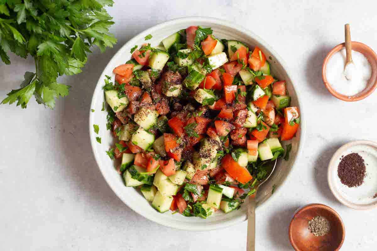 mediterranean cucumber tomato salad in a bowl with a spoon topped with sumac next to a bunch of parsley, and small bowls of olive oil salt, pepper and sumac.