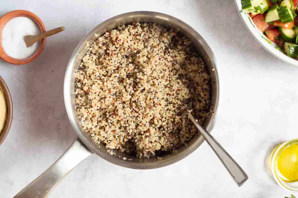 cooked quinoa in a saucepan with a spoon next to a bowl of salt, a bowl of olive oil and the mediterranean cucumber tomato salad.