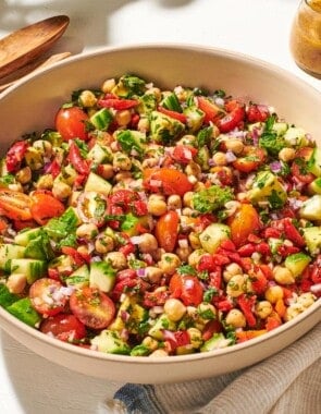 a close up of vegan chickpea salad in a serving bowl next to a wooden serving spoon and fork, a jar of dijon dressing, and a cloth napkin.