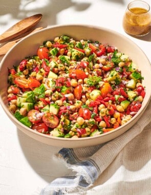 a close up of vegan chickpea salad in a serving bowl next to a wooden serving spoon and fork, a jar of dijon dressing, and a cloth napkin.