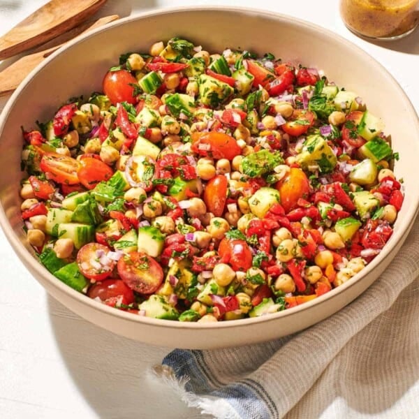 a close up of vegan chickpea salad in a serving bowl next to a wooden serving spoon and fork, a jar of dijon dressing, and a cloth napkin.