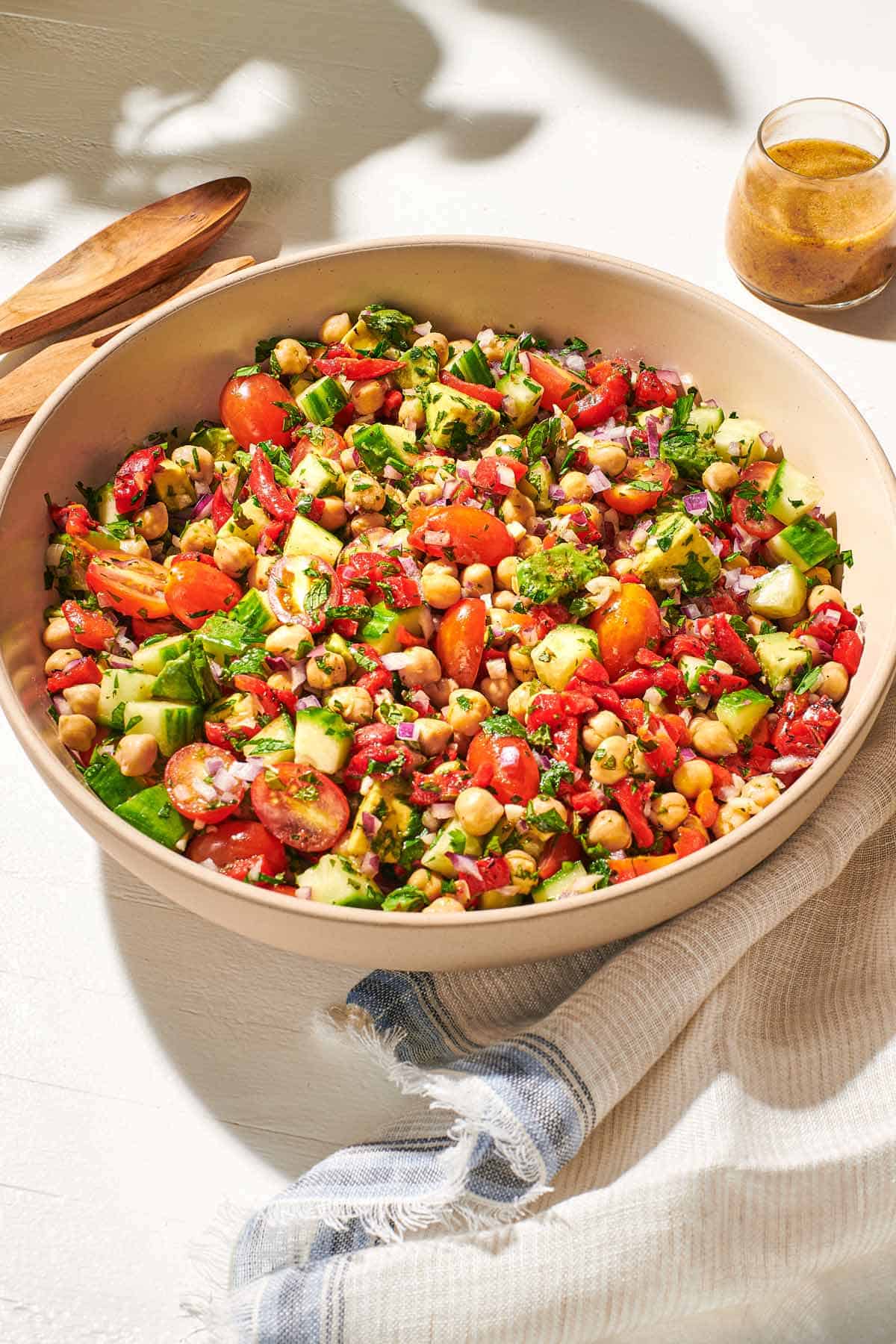 a close up of vegan chickpea salad in a serving bowl next to a wooden serving spoon and fork, a jar of dijon dressing, and a cloth napkin.