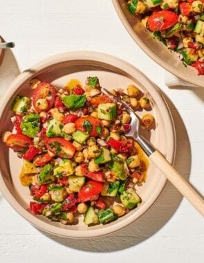 an overhead photo of a bowl of vegan chickpea salad with a fork next to a serving bowl of the salad, 2 more empty bowls and a fork, a jar of dijon dressing with a spoon, and a glass of water.