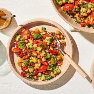an overhead photo of a bowl of vegan chickpea salad with a fork next to a serving bowl of the salad, 2 more empty bowls and a fork, a jar of dijon dressing with a spoon, and a glass of water.